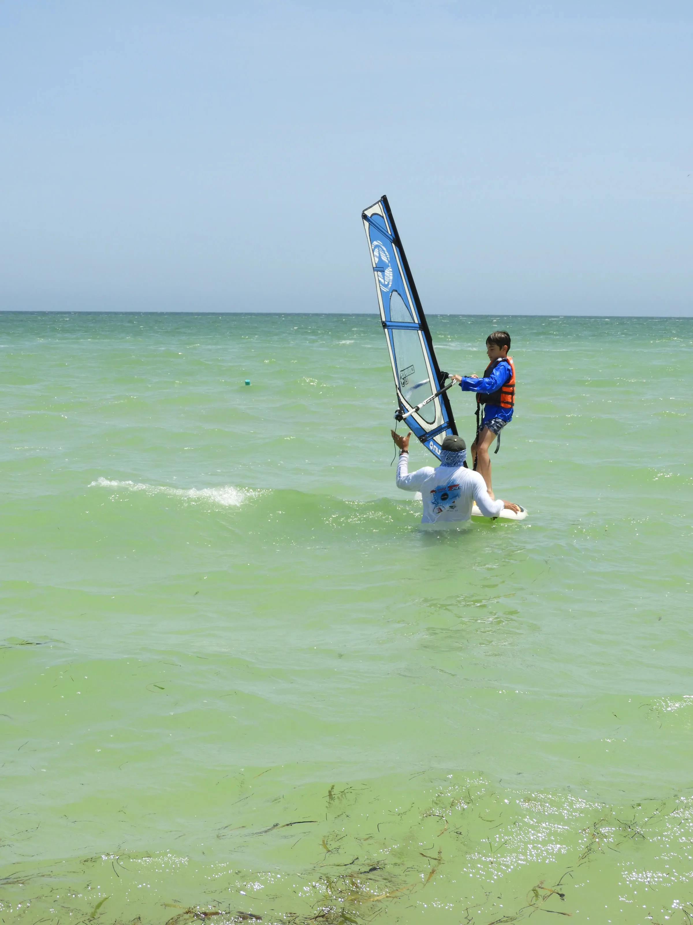 Instructor de pie en el agua ayuda a un niño con chaleco salvavidas a sostener la botavara de una tabla de windsurf.