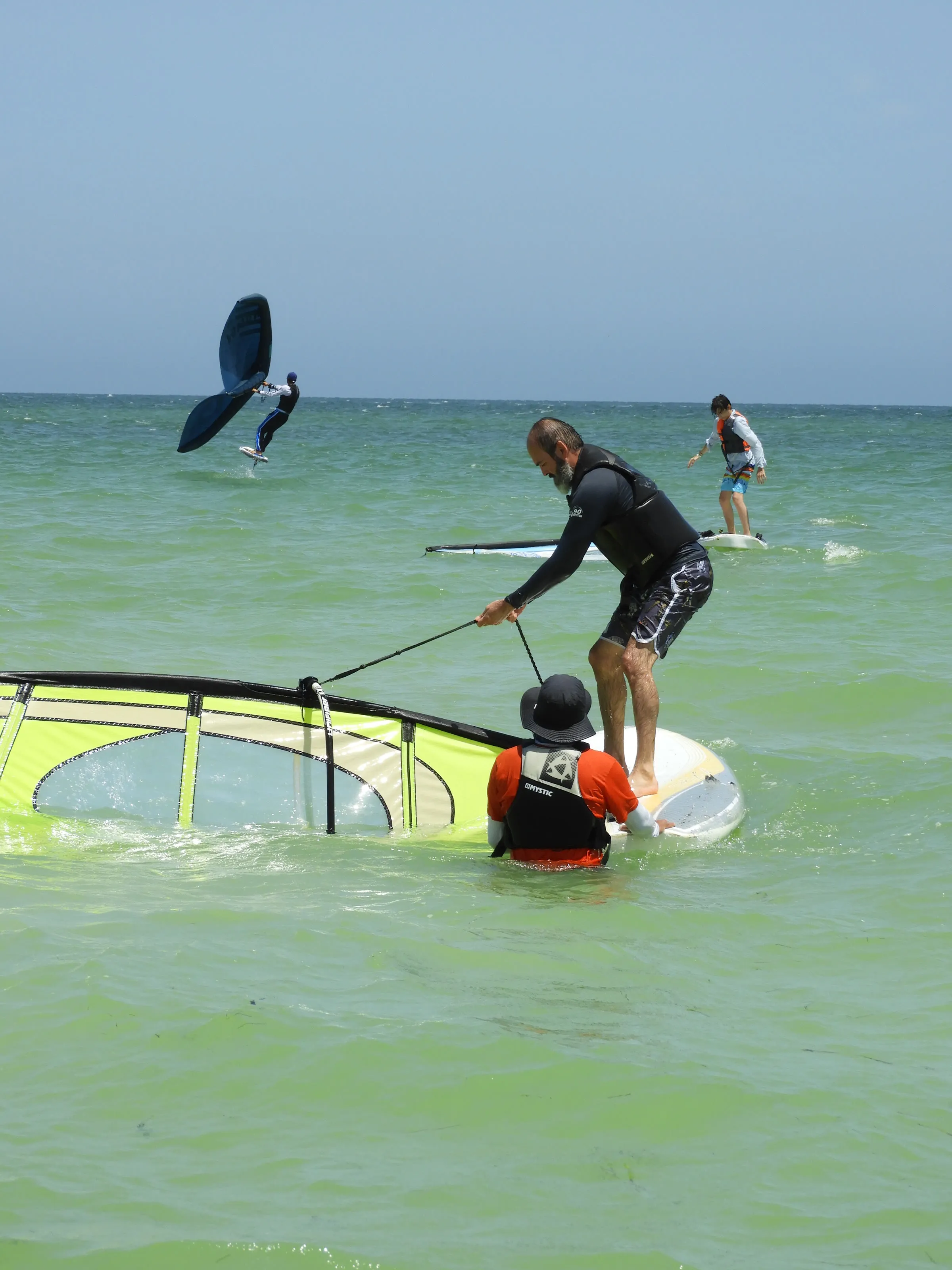 Instructor endereza la vela caída de una tabla de windsurf junto a un niño con chaleco; al fondo, un practicante de wing foil salta sobre el agua.