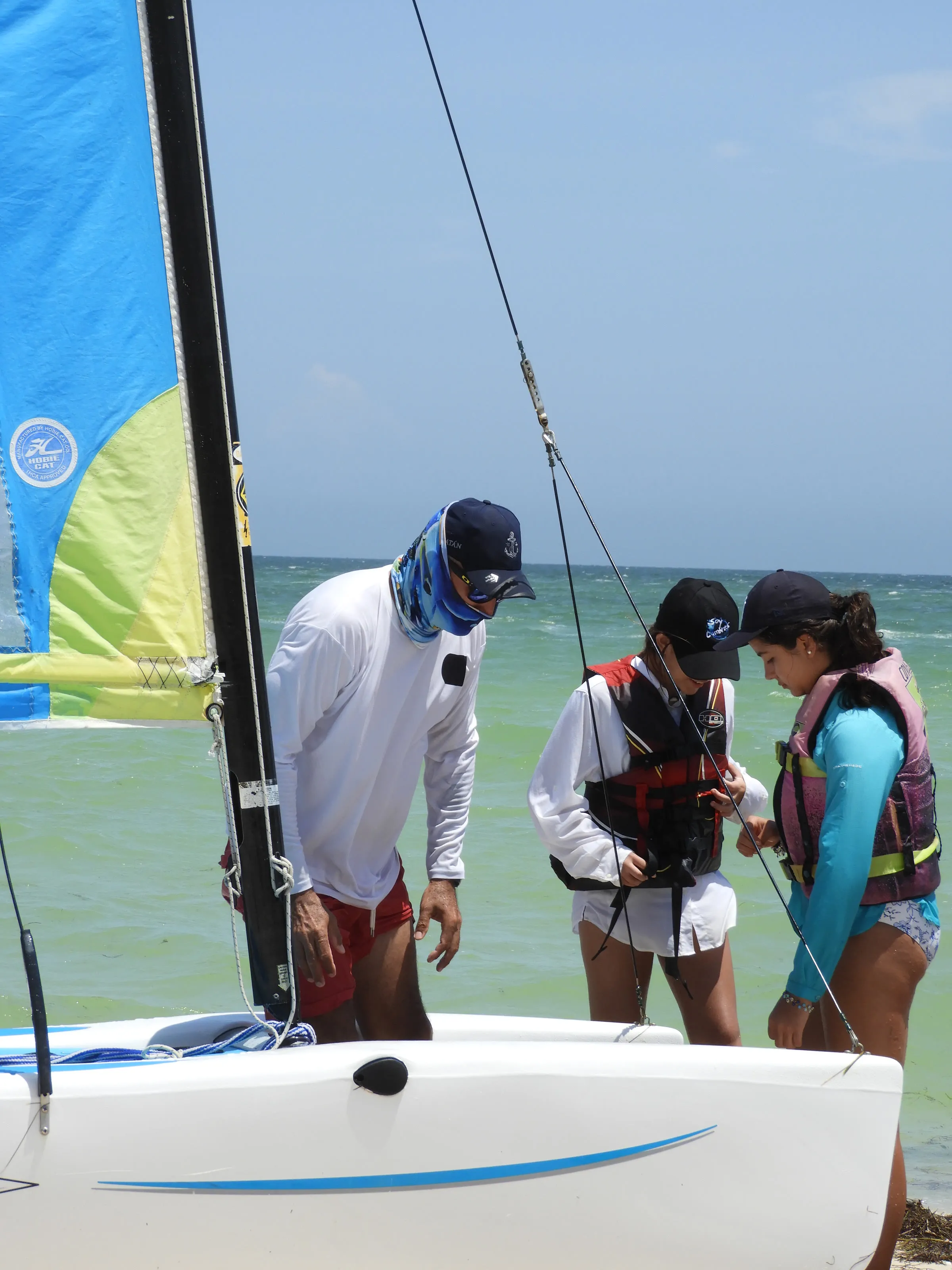 Instructor ayuda a dos estudiantes a ajustarse el chaleco salvavidas junto a un Hobie Cat con la vela izada en la playa.