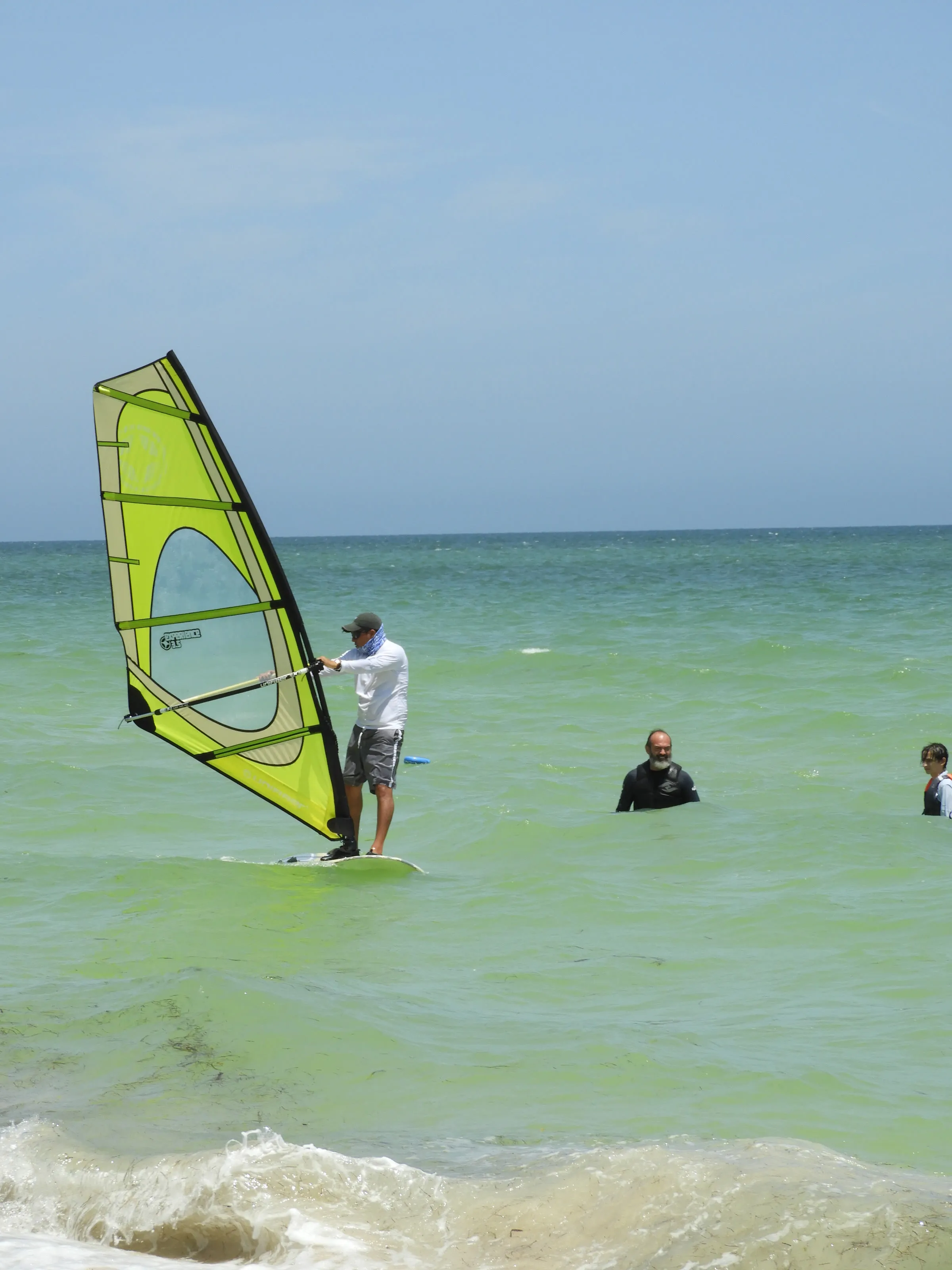 Hombre con gorra, lentes de sol y paliacate cubriéndole el rostro navega de pie en una tabla de windsurf con vela verde; detrás, un hombre con barba y canas nada en el agua, y a un lado un niño observa.