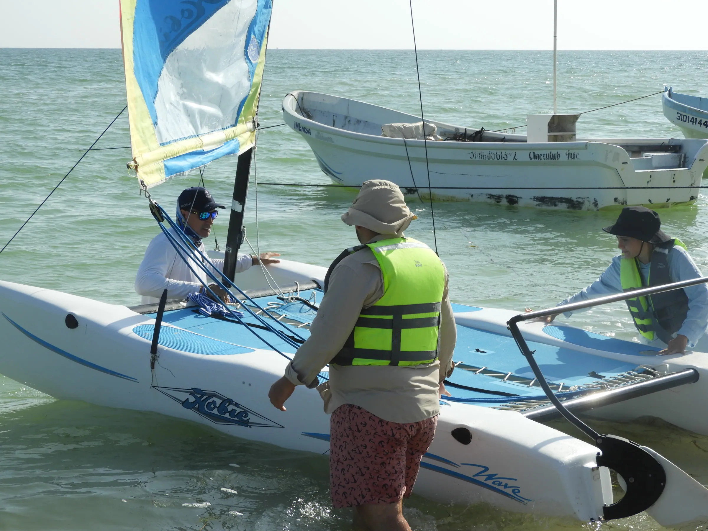 Tres personas suben a un Hobie Wave con la vela izada, junto a lanchas de pesca ancladas.