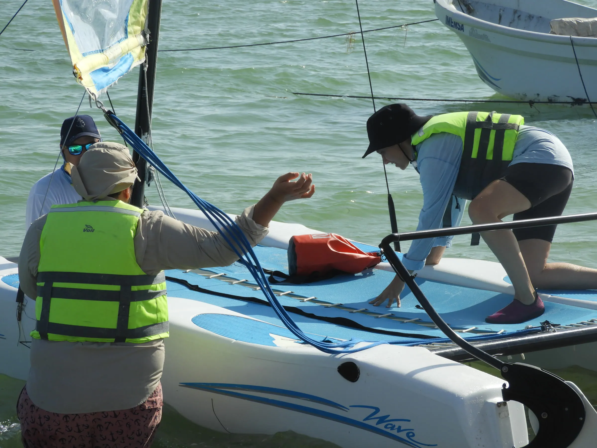 Dos personas con chaleco salvavidas se apoyan sobre la red de un Hobie Wave mientras un tercero observa de pie en el agua.