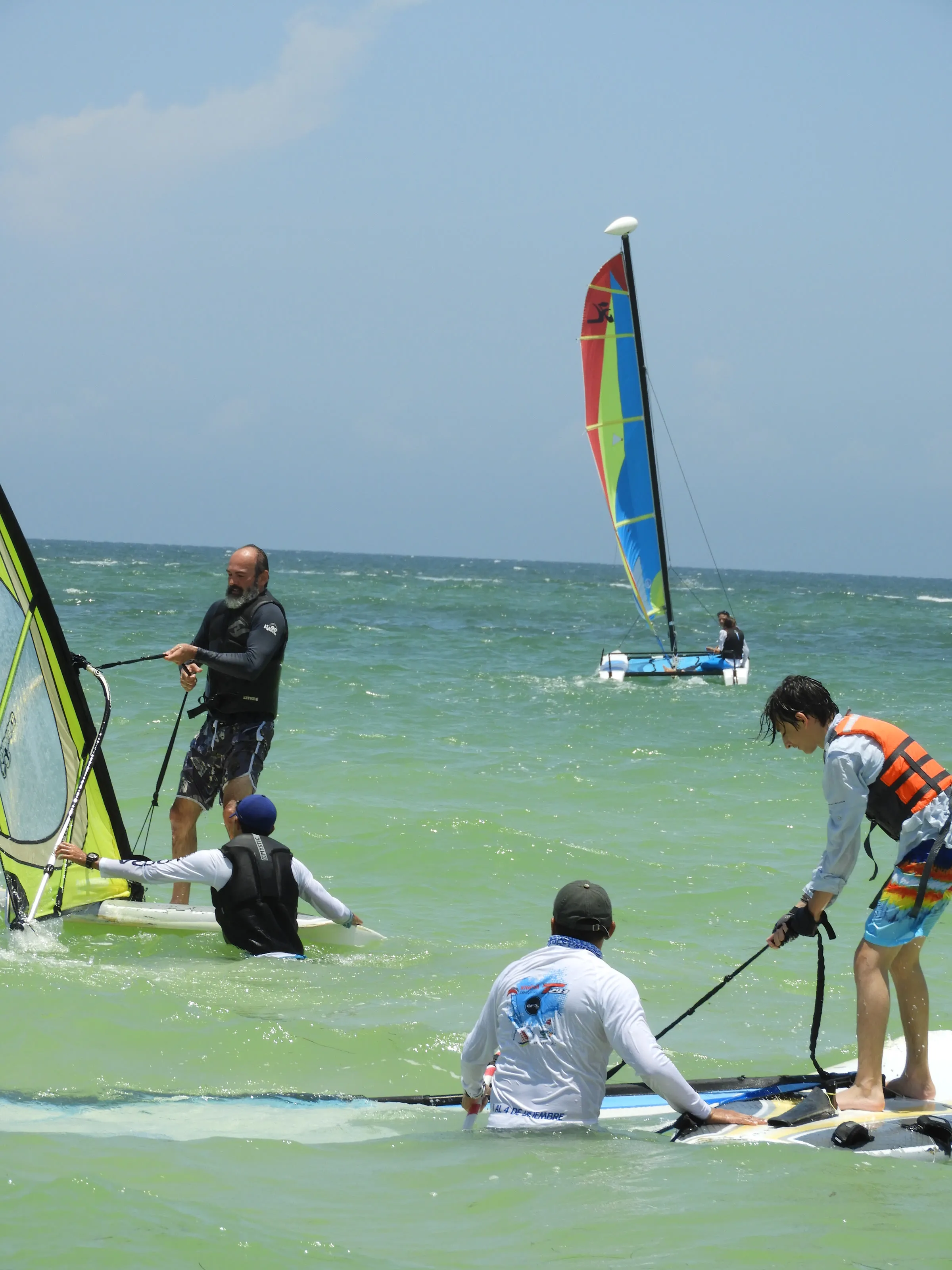 Instructor y dos niños con tablas de windsurf en primer plano; al fondo navega un Hobie Cat con vela azul, roja y verde.