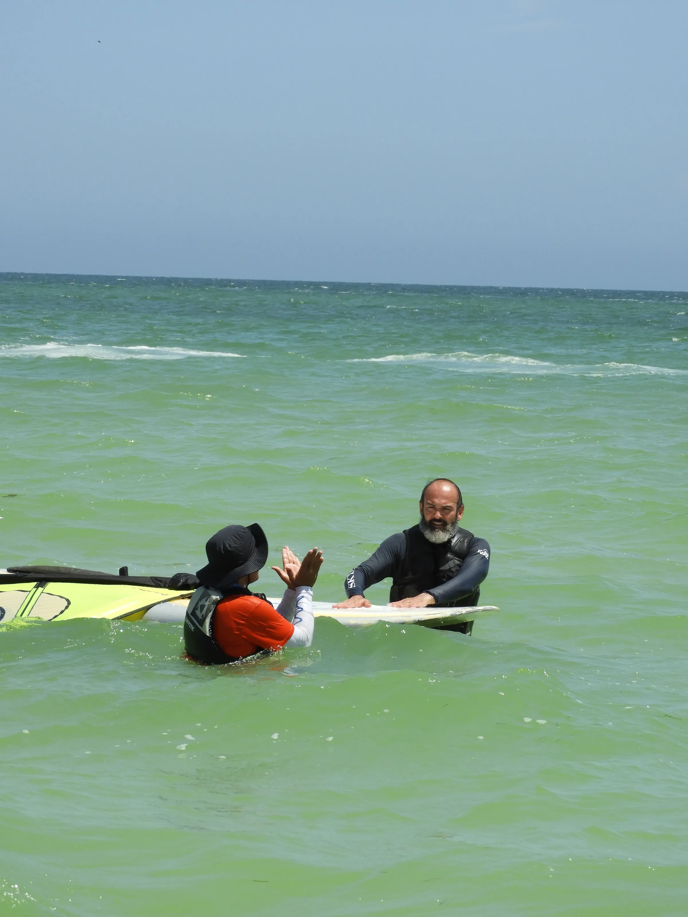 Instructor con barba choca la mano con un niño, ambos en el agua junto a una tabla de windsurf.