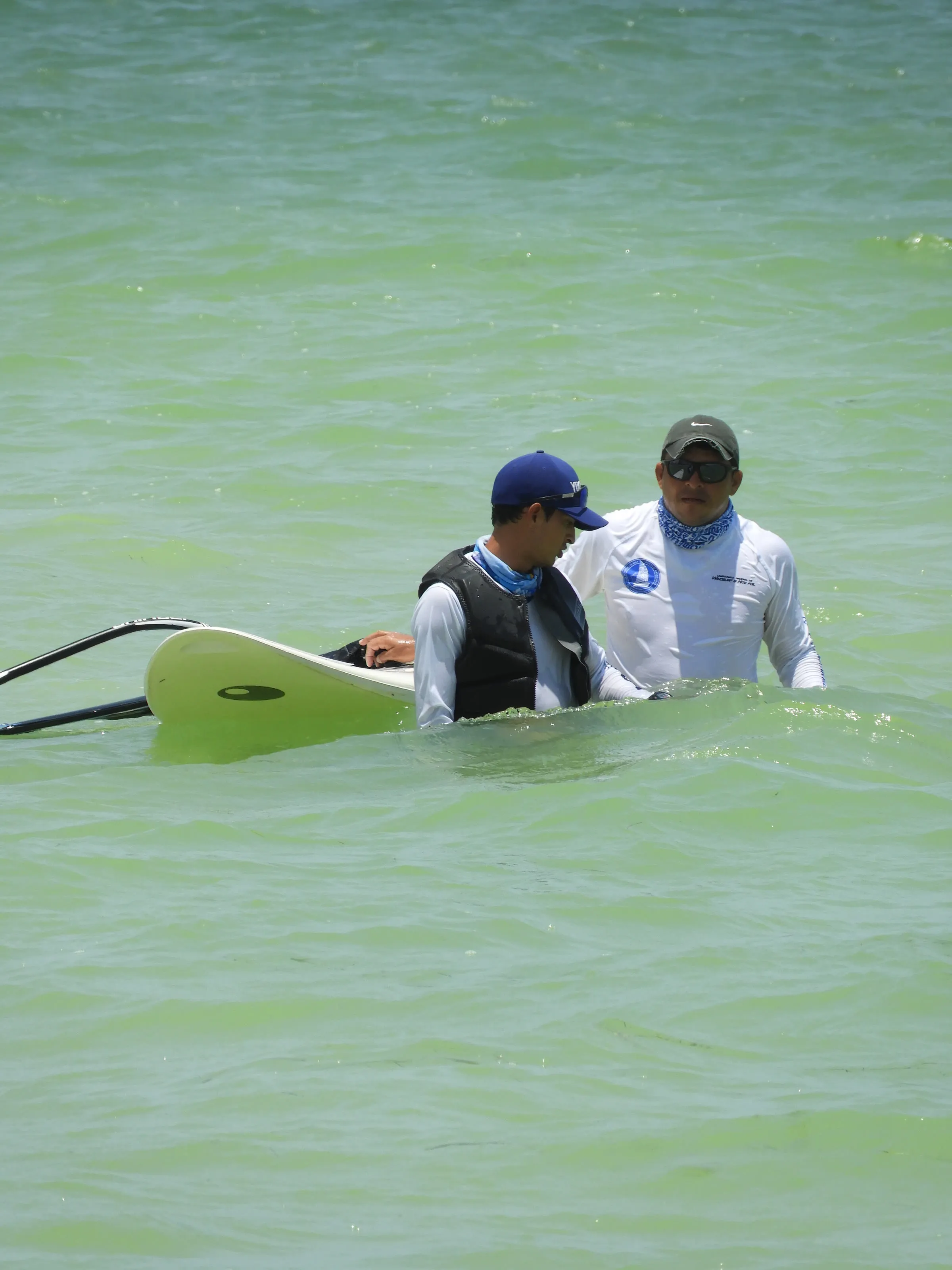 Dos instructores de pie en el agua conversan junto a una tabla de windsurf con vela amarilla.