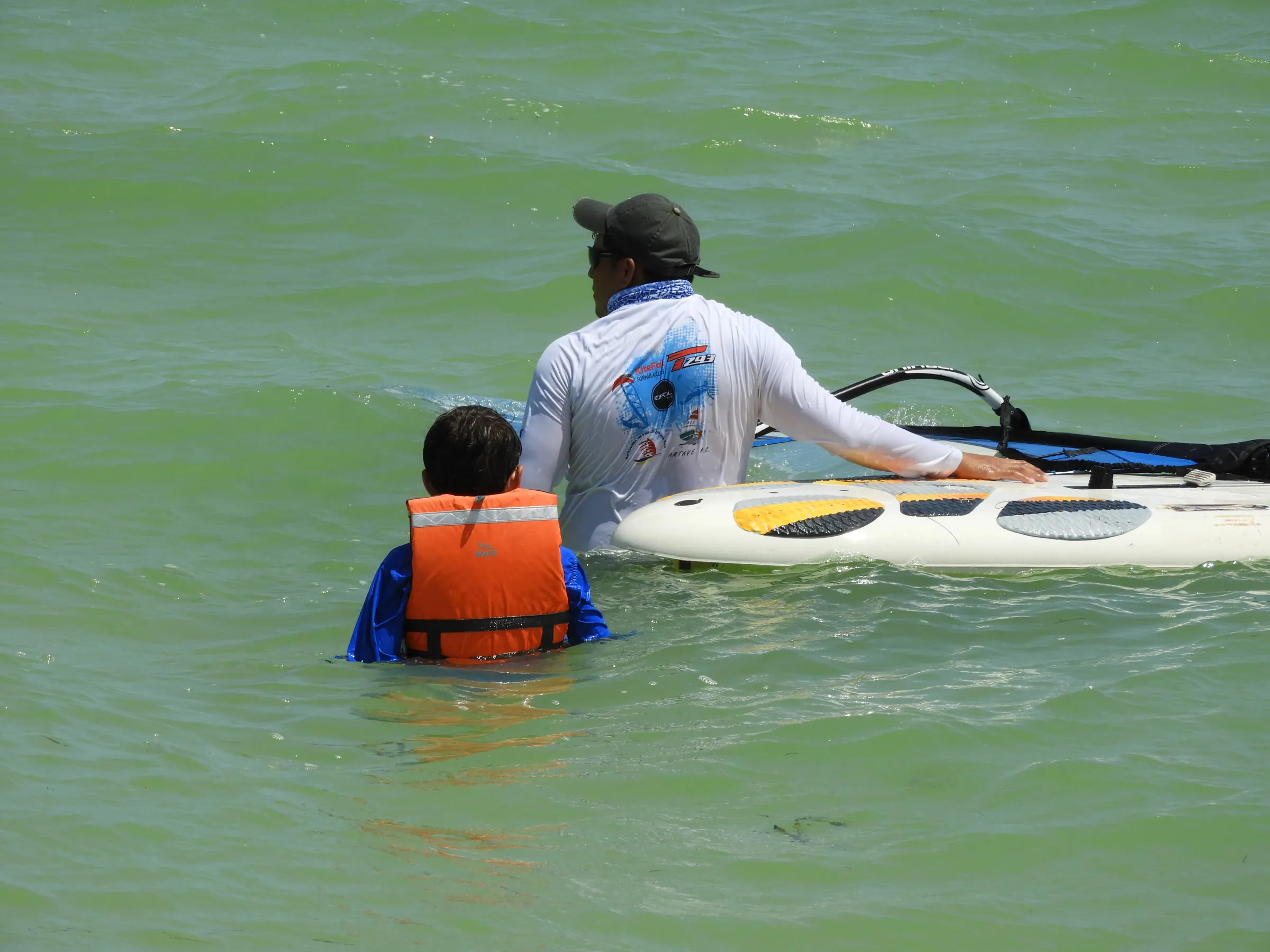 Instructor y un niño con chaleco salvavidas naranja, vistos de espaldas, junto a una tabla de windsurf.