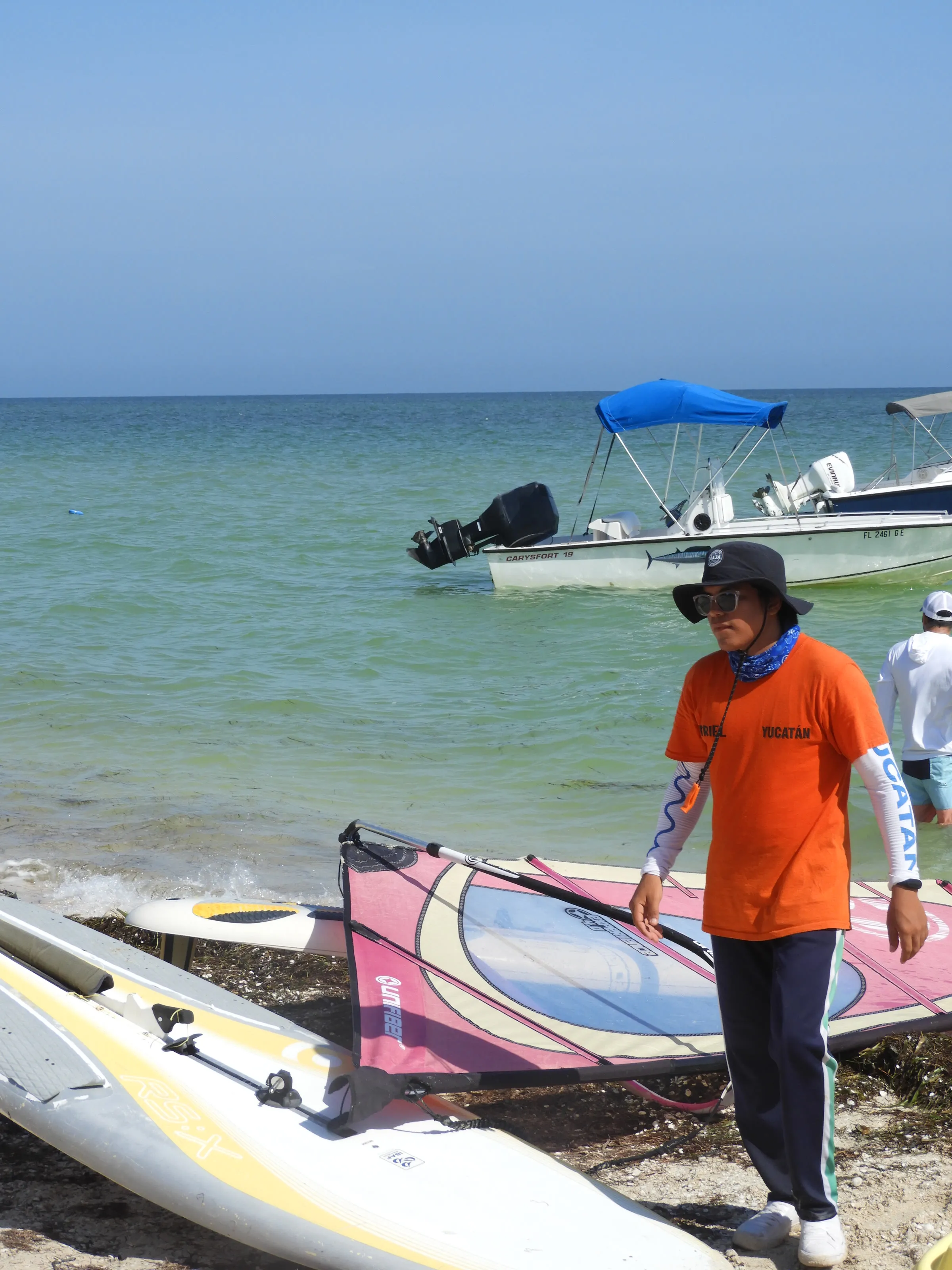 Instructor con sombrero, lentes de sol y camiseta naranja de Yucatán de pie junto a tablas y velas de windsurf tendidas en la arena; al fondo, una lancha con toldo azul anclada en el mar.