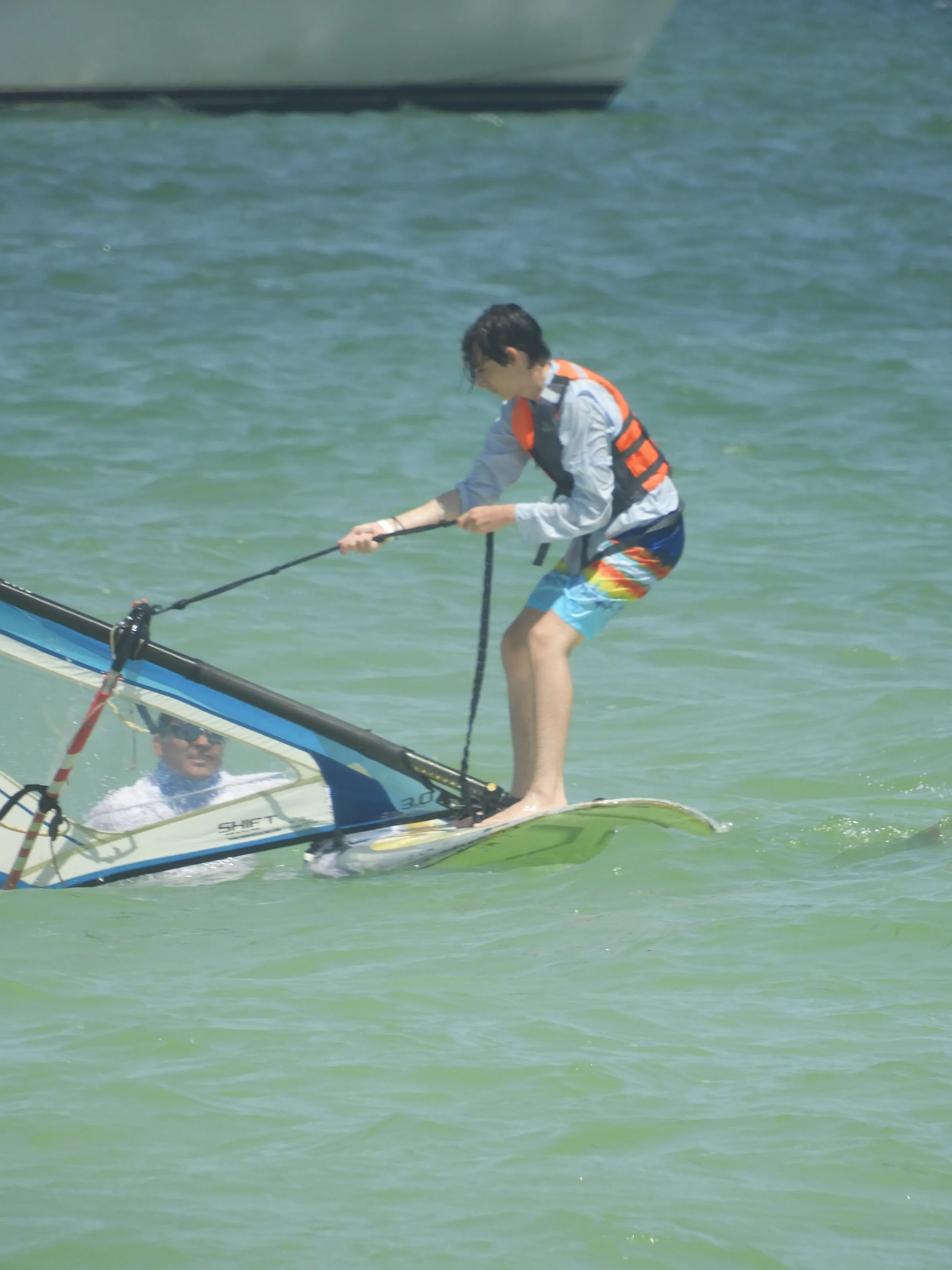 Niño con chaleco salvavidas naranja de pie sobre una tabla de windsurf; un instructor lo observa desde el agua.