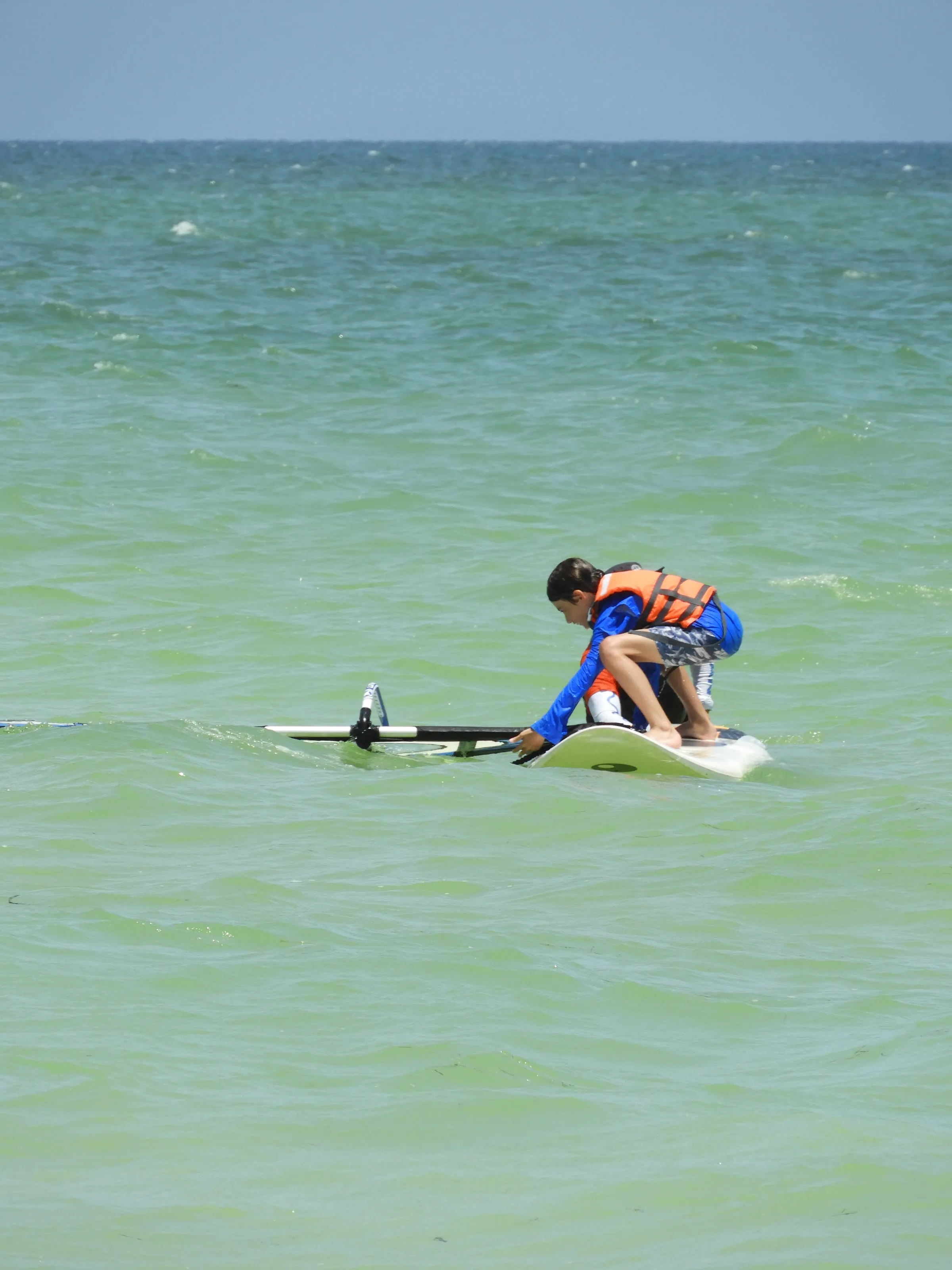 Niño con chaleco salvavidas naranja agachado sobre una tabla de windsurf en el agua.