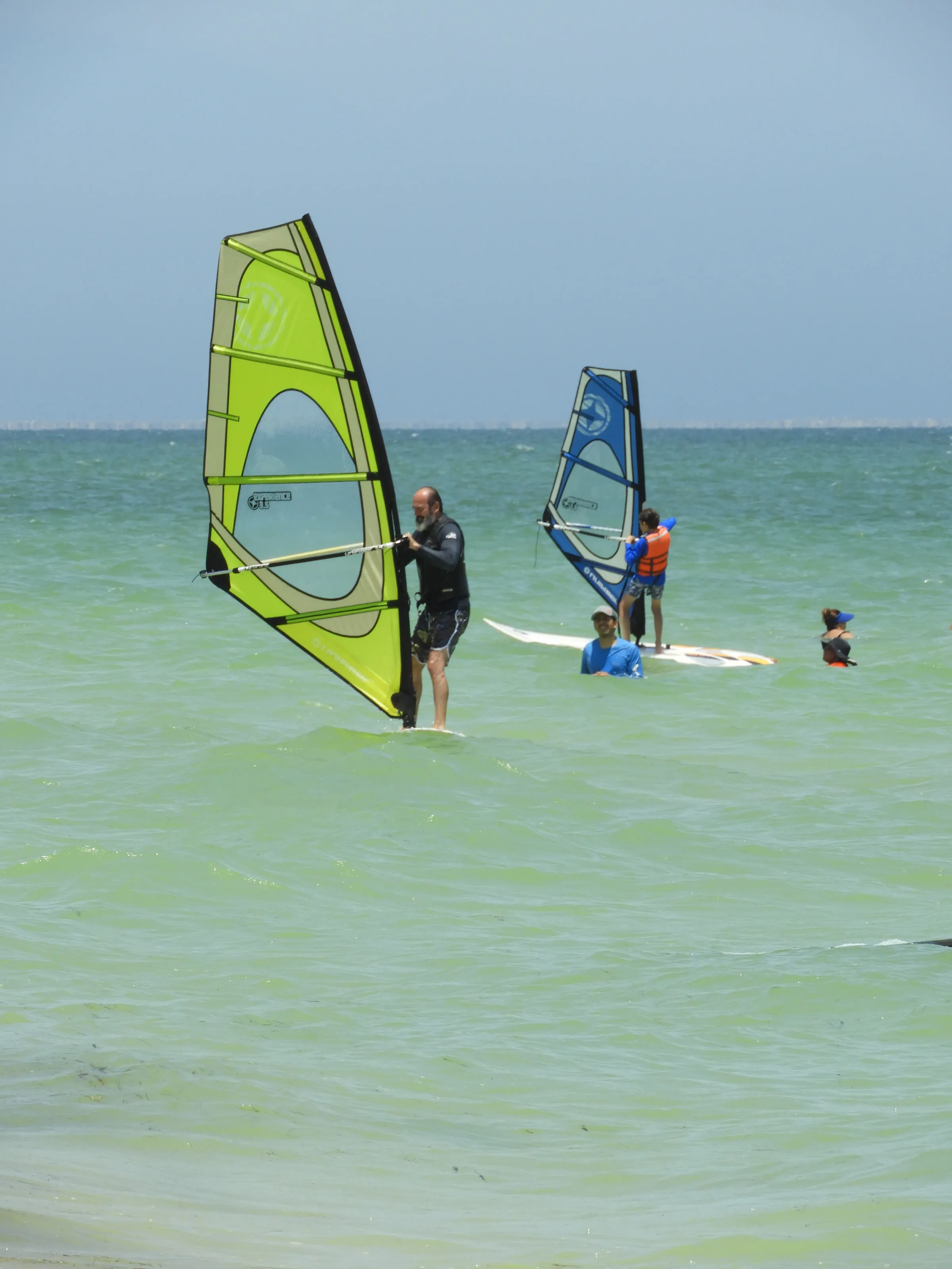 Hombre con barba navega de pie en una tabla de windsurf con vela verde; detrás, un niño con chaleco naranja practica con otra vela junto a dos personas más en el agua.