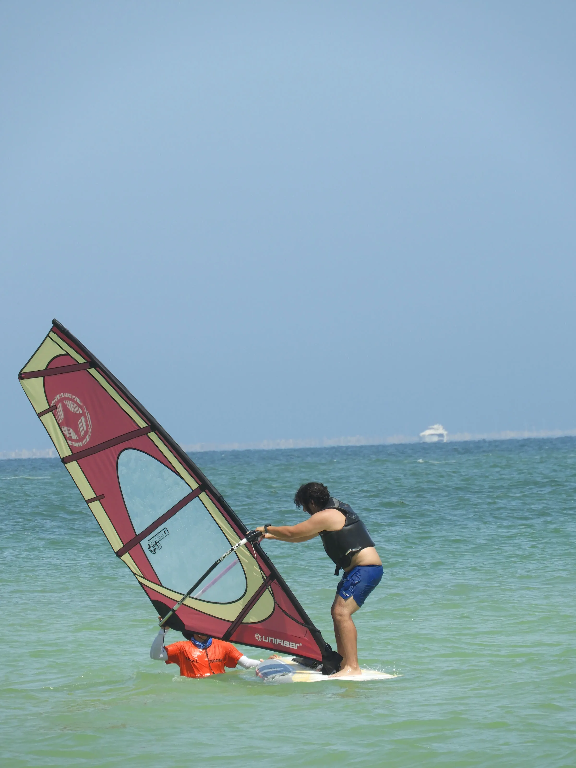 Hombre navega de pie en una tabla de windsurf con vela roja y crema; un instructor con camiseta naranja sostiene la tabla desde el agua.