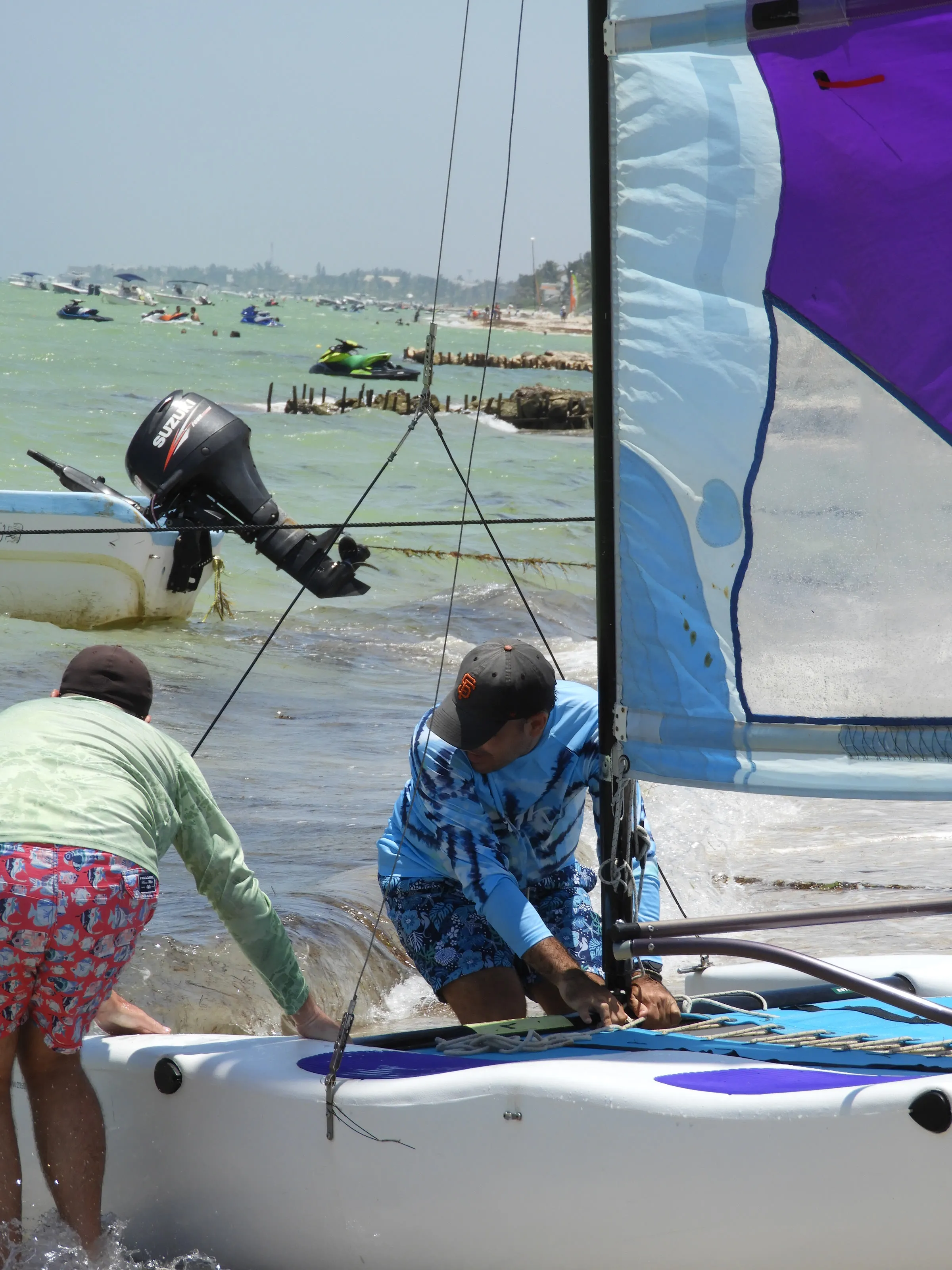 Dos hombres preparan un Hobie Cat con vela azul y morada en la orilla; al fondo, lanchas y motos acuáticas en la playa.