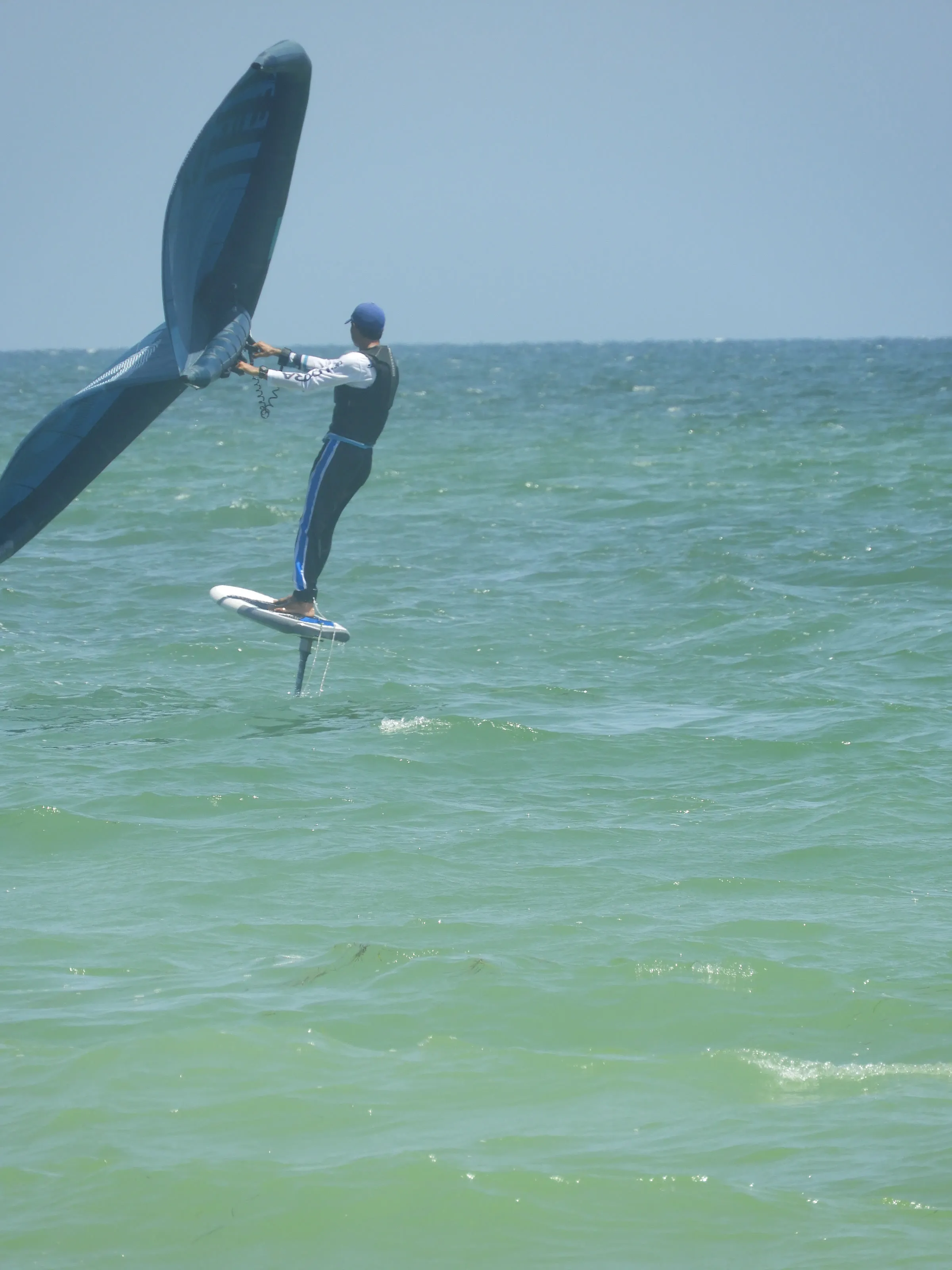 Practicante de wing foil eleva la tabla sobre el agua sostenido por el hidroala, con el ala inflable en las manos.